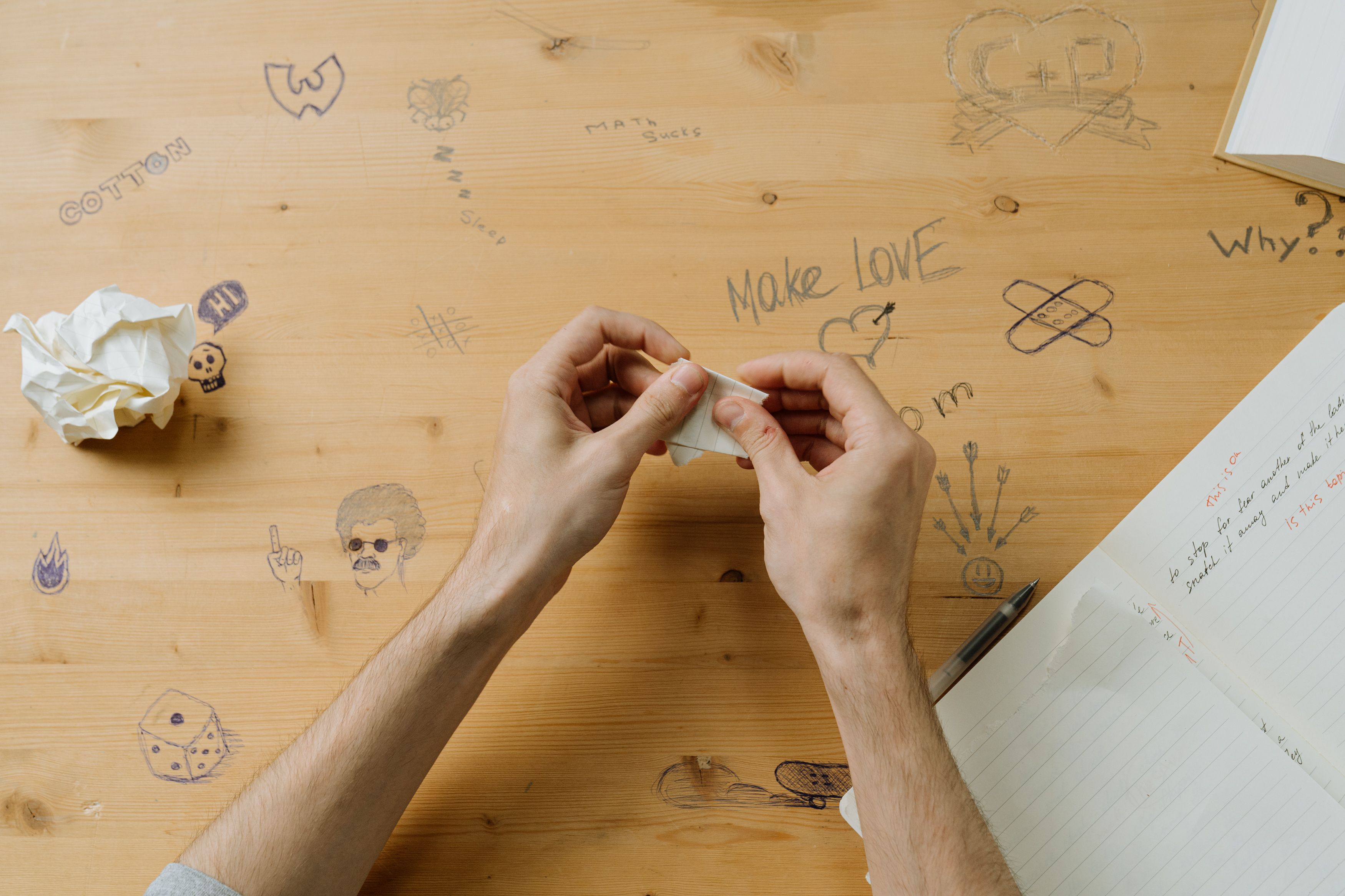 A top-down view of a desk with doodles, hands folding paper, and a notebook.