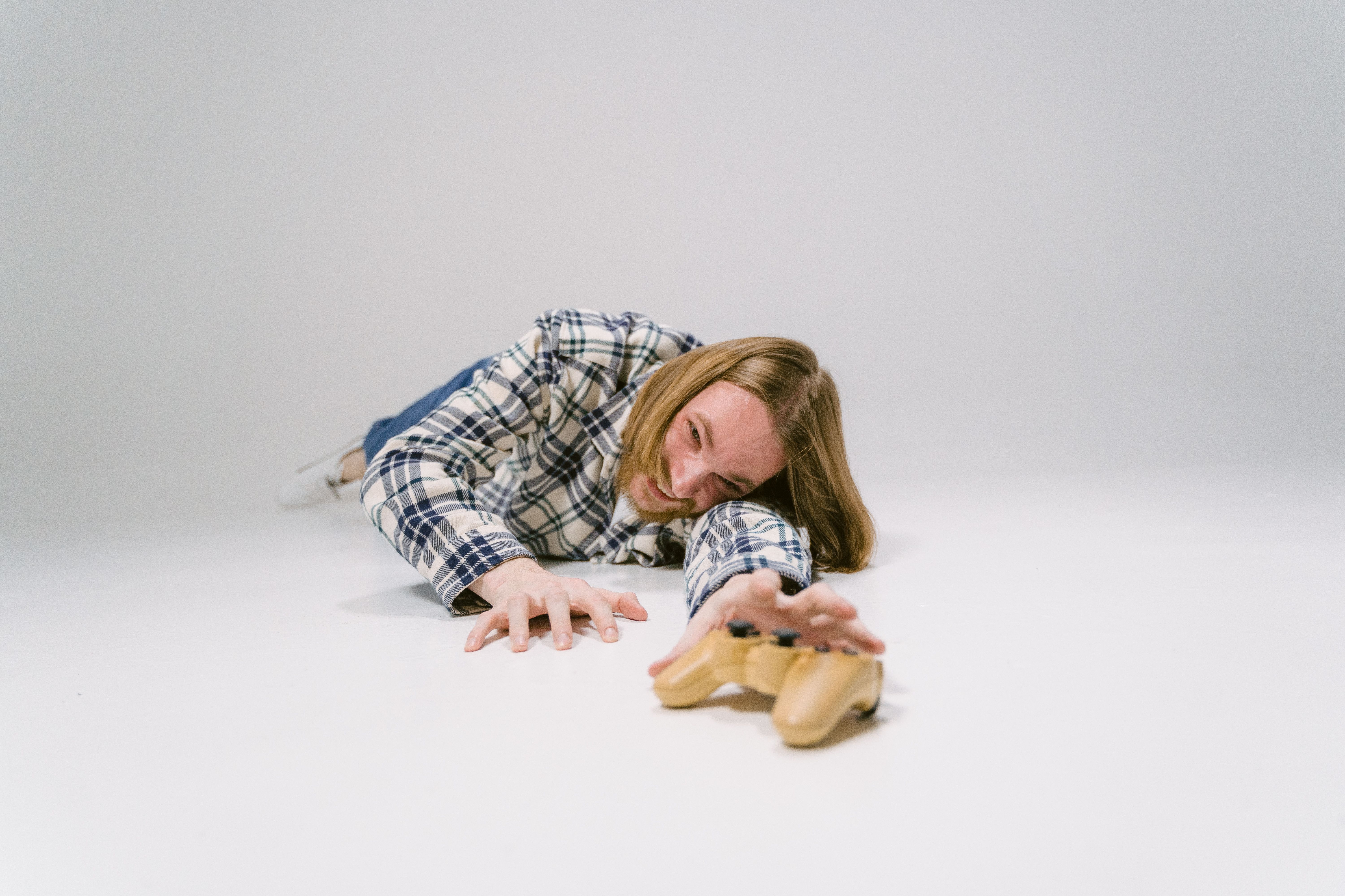 Young man in plaid shirt reaching for a controller on white studio floor.