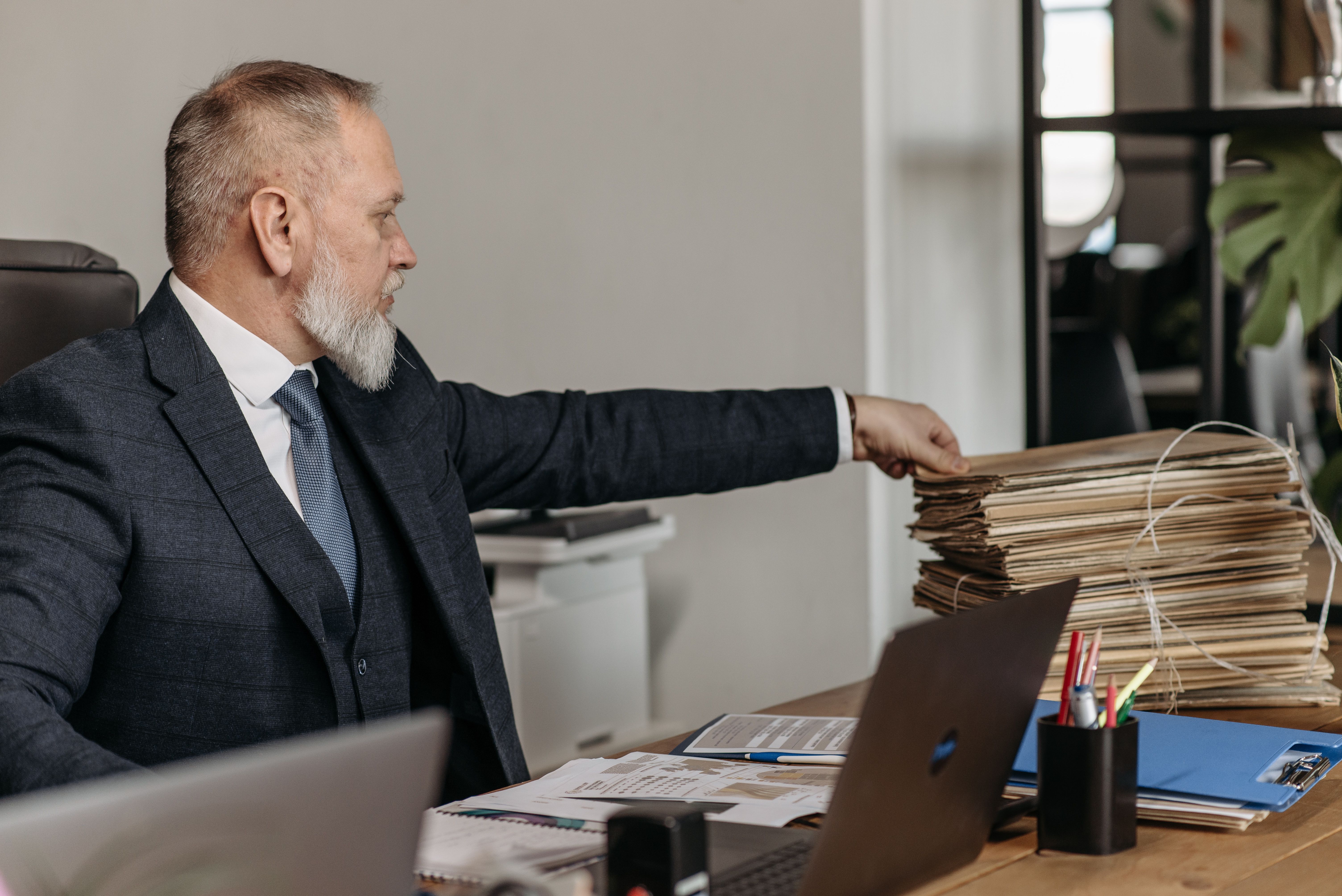 Mature businessman reaching for a stack of files on his desk in a modern office environment.