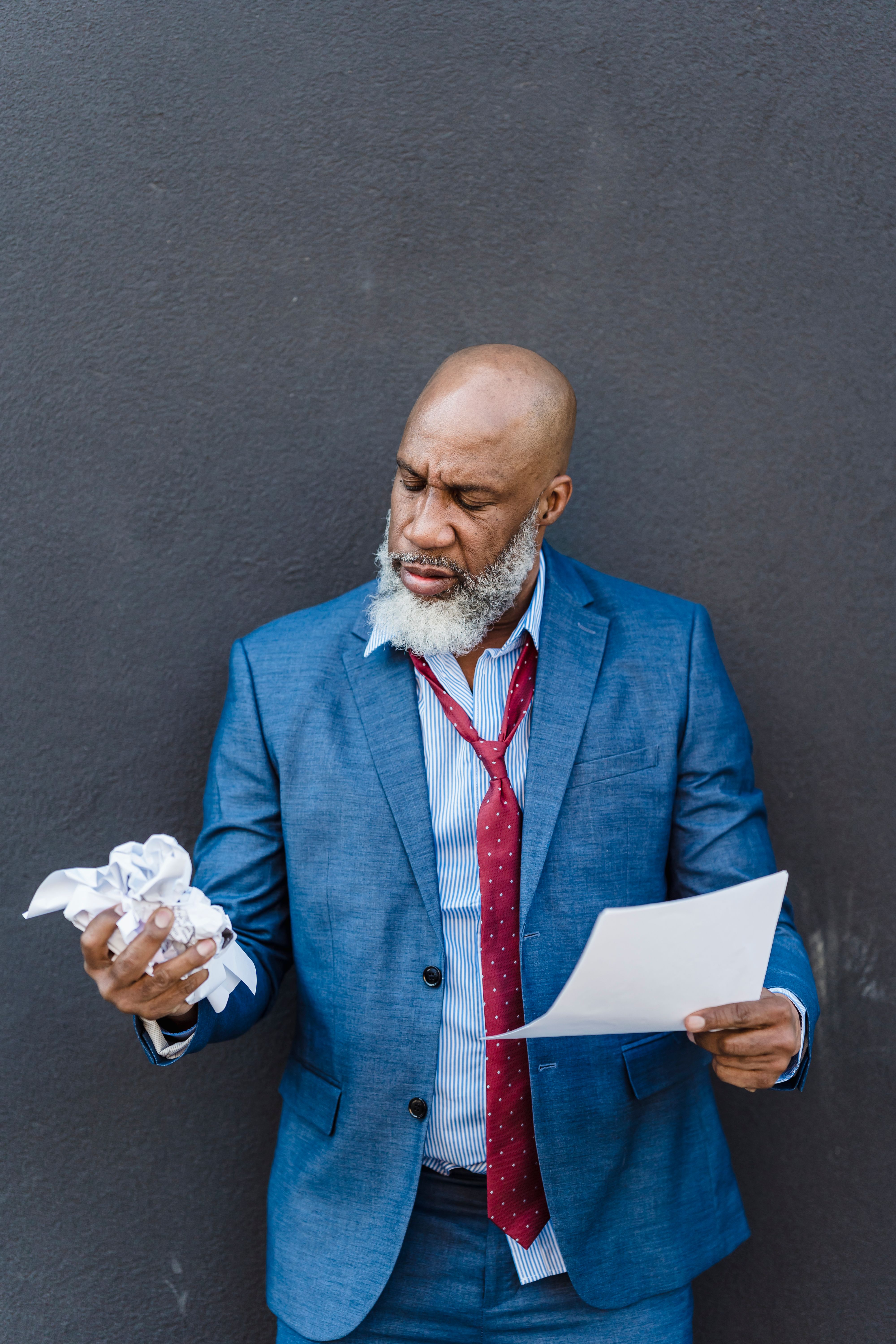 Unhappy African American male entrepreneur in formal suit making crumple of official papers on gray background on street in city