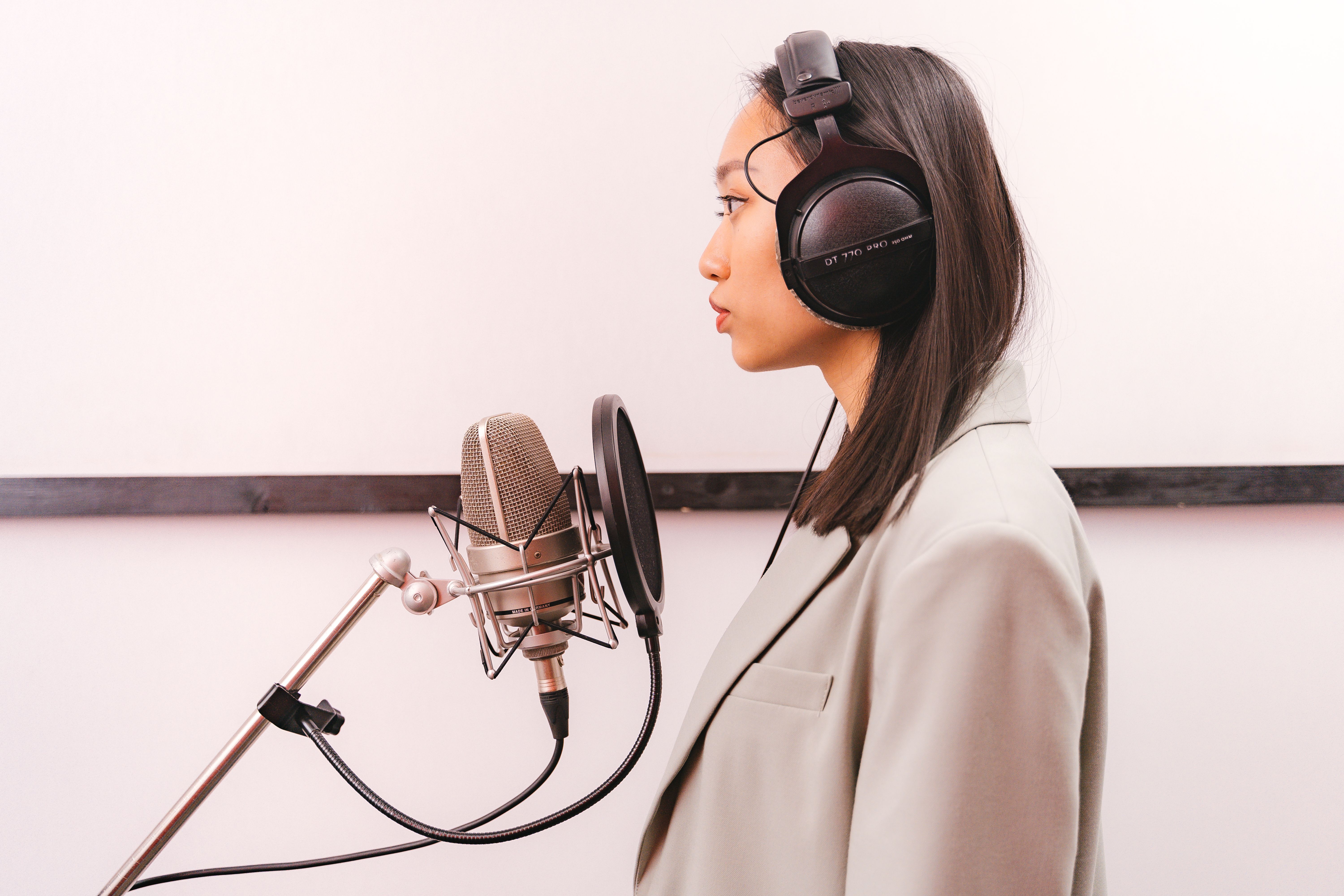 Side view of an Asian woman with headphones singing into a condenser microphone in a studio.